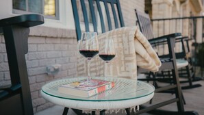 Una mesa de vidrio con dos copas de vino y un libro, sobre un balcón con una silla y una manta.