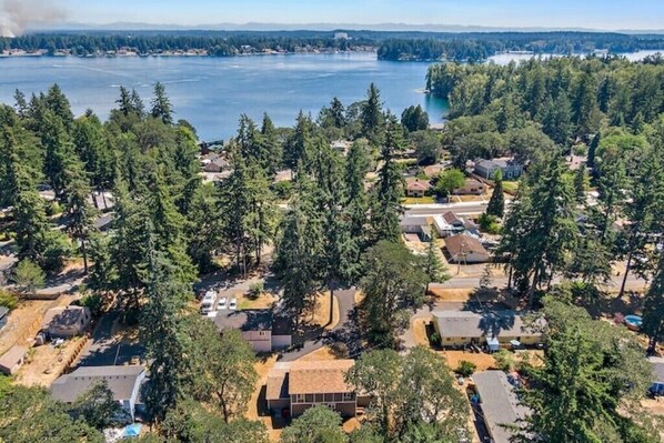 An aerial view of a residential area with houses surrounded by dense forest, a lake in the background, and a clear sky.