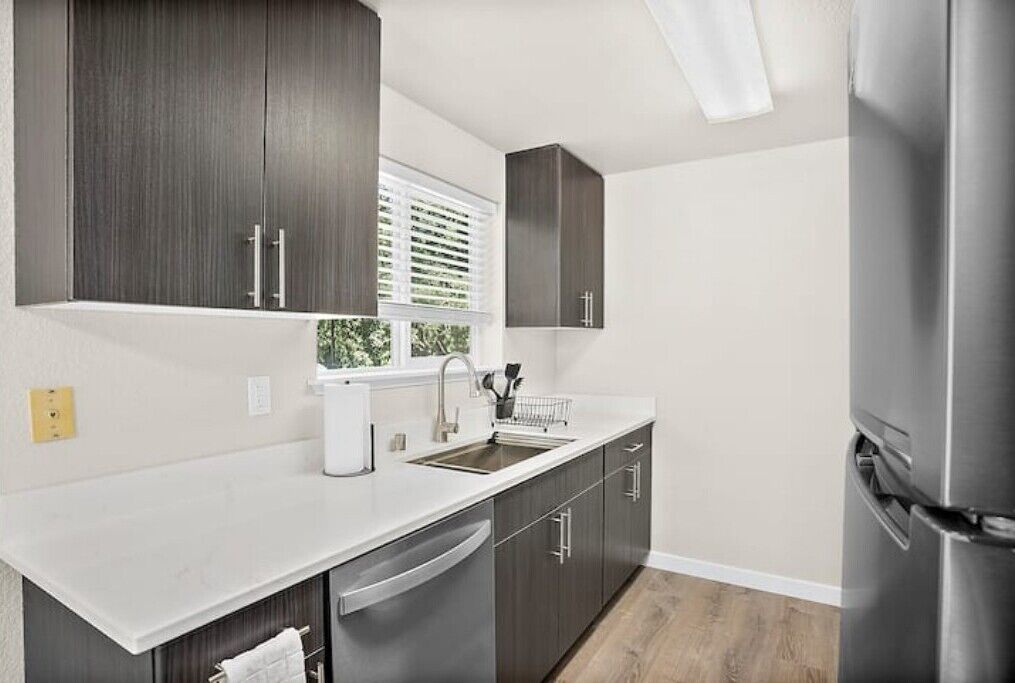 A modern kitchen with dark wood cabinets, white countertops, stainless steel appliances, and a window with blinds.