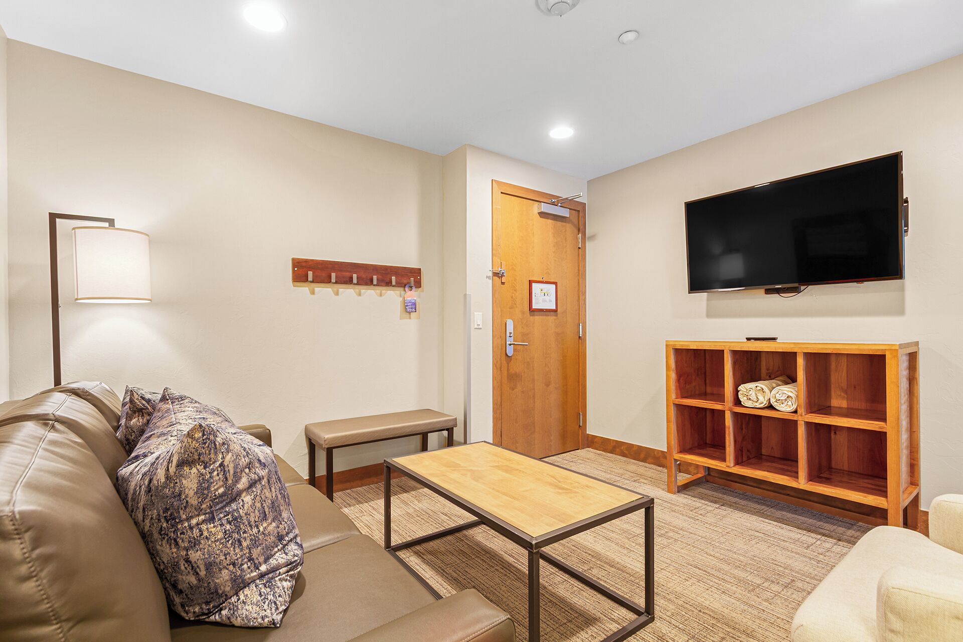 Uma sala de estar com sofá, mesa de centro, televisão, estante de madeira e porta com cabideiro.