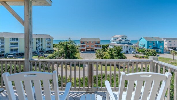 A wooden deck with white chairs overlooking a beachfront property with colorful buildings and a clear blue sky. - Ocean Views and Pool, King Bed, Beach Access at end of Driveway! (Emerald Isle)