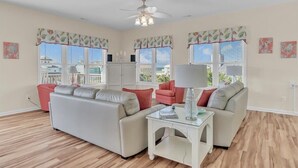 A living room with a beige sofa, a red armchair, and a glass coffee table. There are two windows with curtains, a ceiling fan, and artwork on the walls. - Ocean Views and Pool, King Bed, Beach Access at end of Driveway! (Emerald Isle)