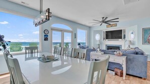 A spacious living area with a large dining table, a sofa, and a TV. There are multiple windows offering a view of the ocean. - 5BR, Ocean Front, 1/2 Duplex! (Emerald Isle)