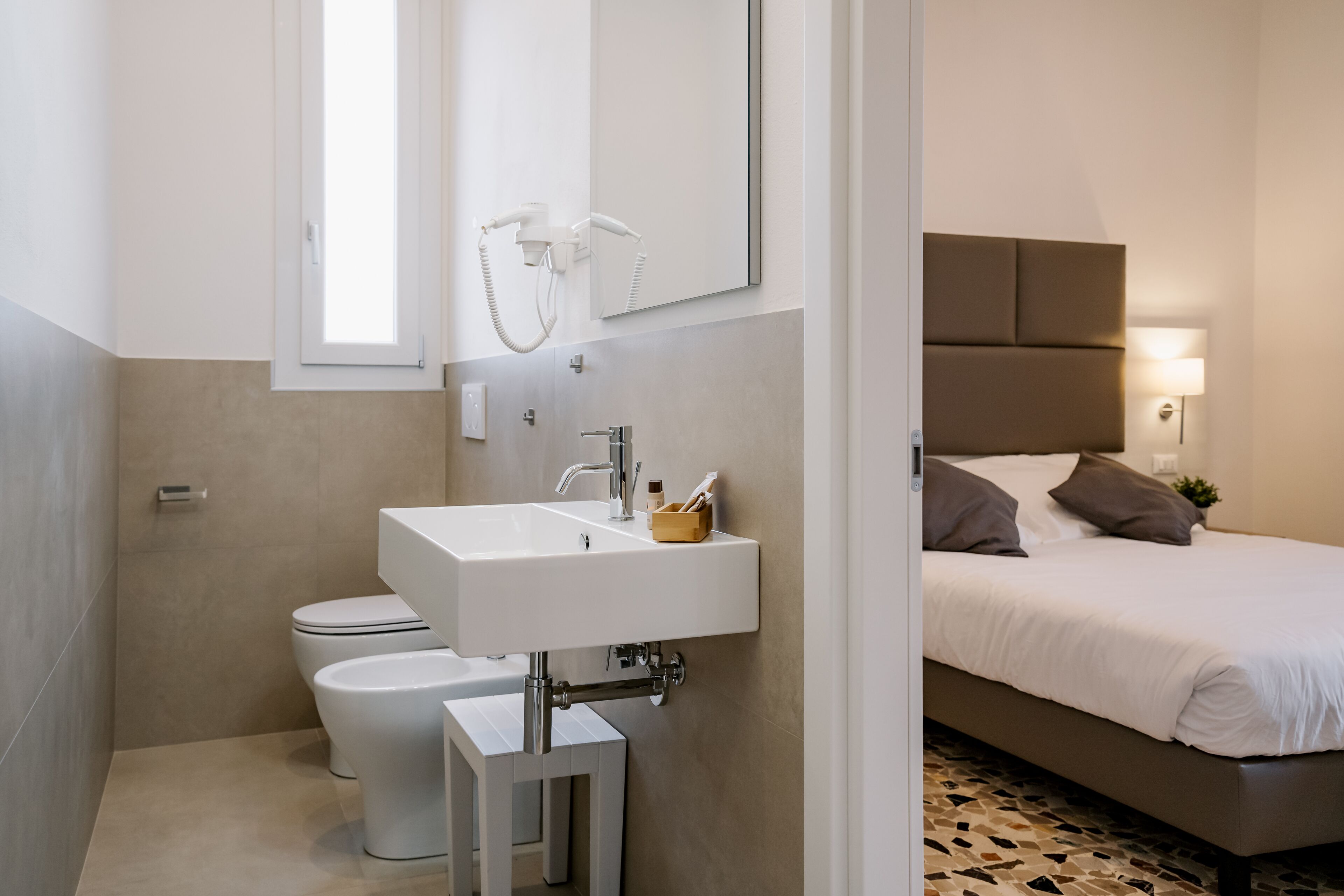 Et moderne badeværelse med en hvid vask, toilet og brusebad. Der er en seng med sengegavl og et natbord med lampe.