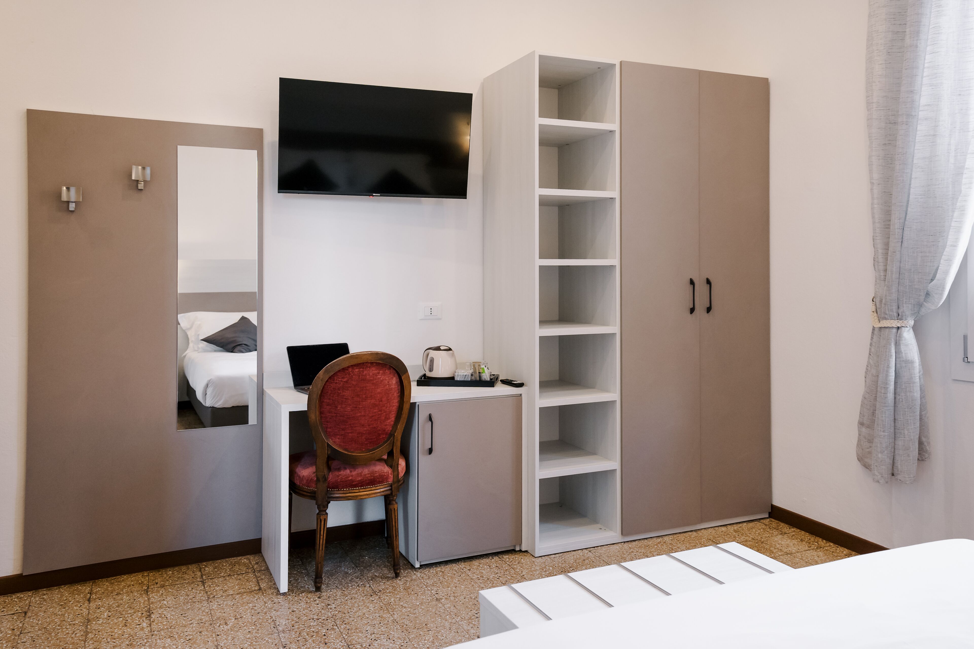 A hotel room with a bed, a desk with a laptop, a TV mounted above the desk, a wardrobe with multiple shelves, and a chair.