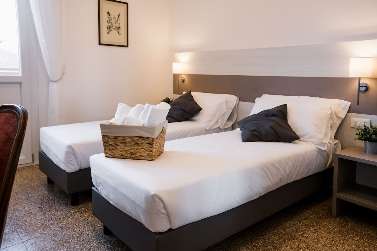 Une chambre d’hôtel avec deux lits simples, un panier en osier sur le lit de gauche et une table de chevet avec une plante.