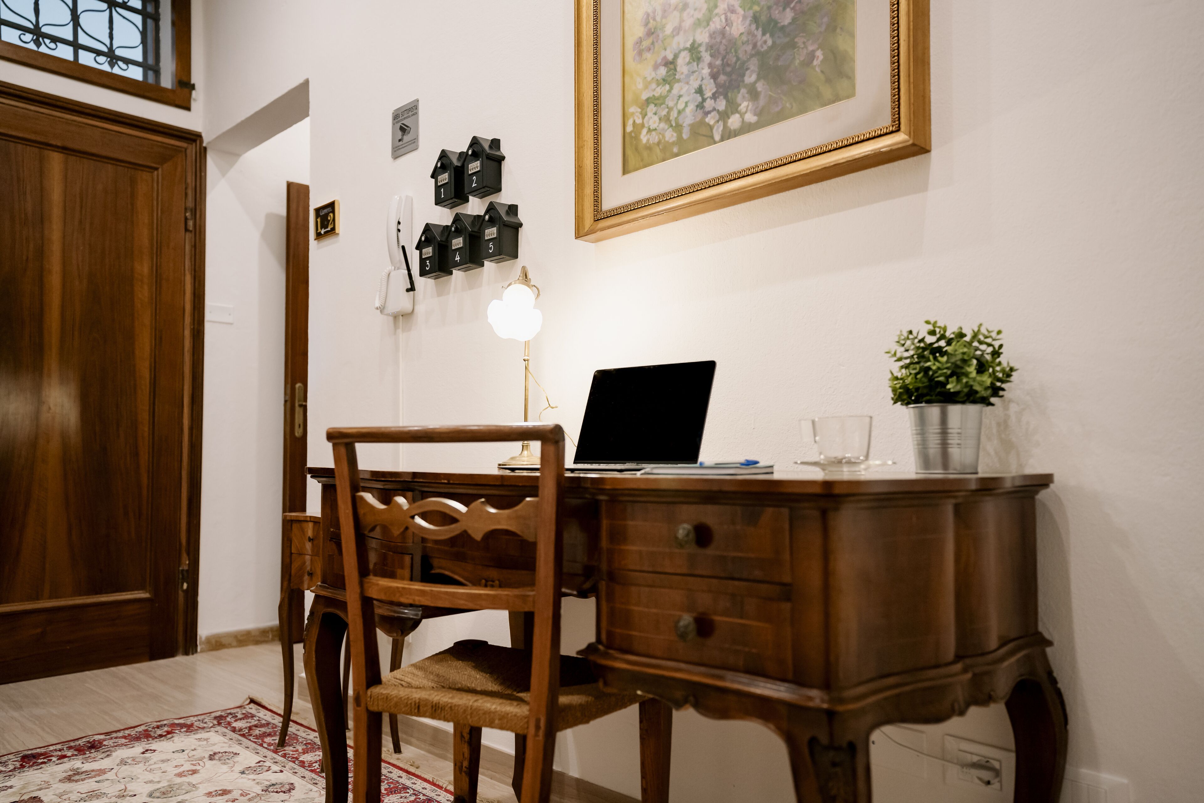 Un escritorio de madera con una computadora portátil, una lámpara y una planta en maceta. Hay una silla frente al escritorio y una puerta a la izquierda.