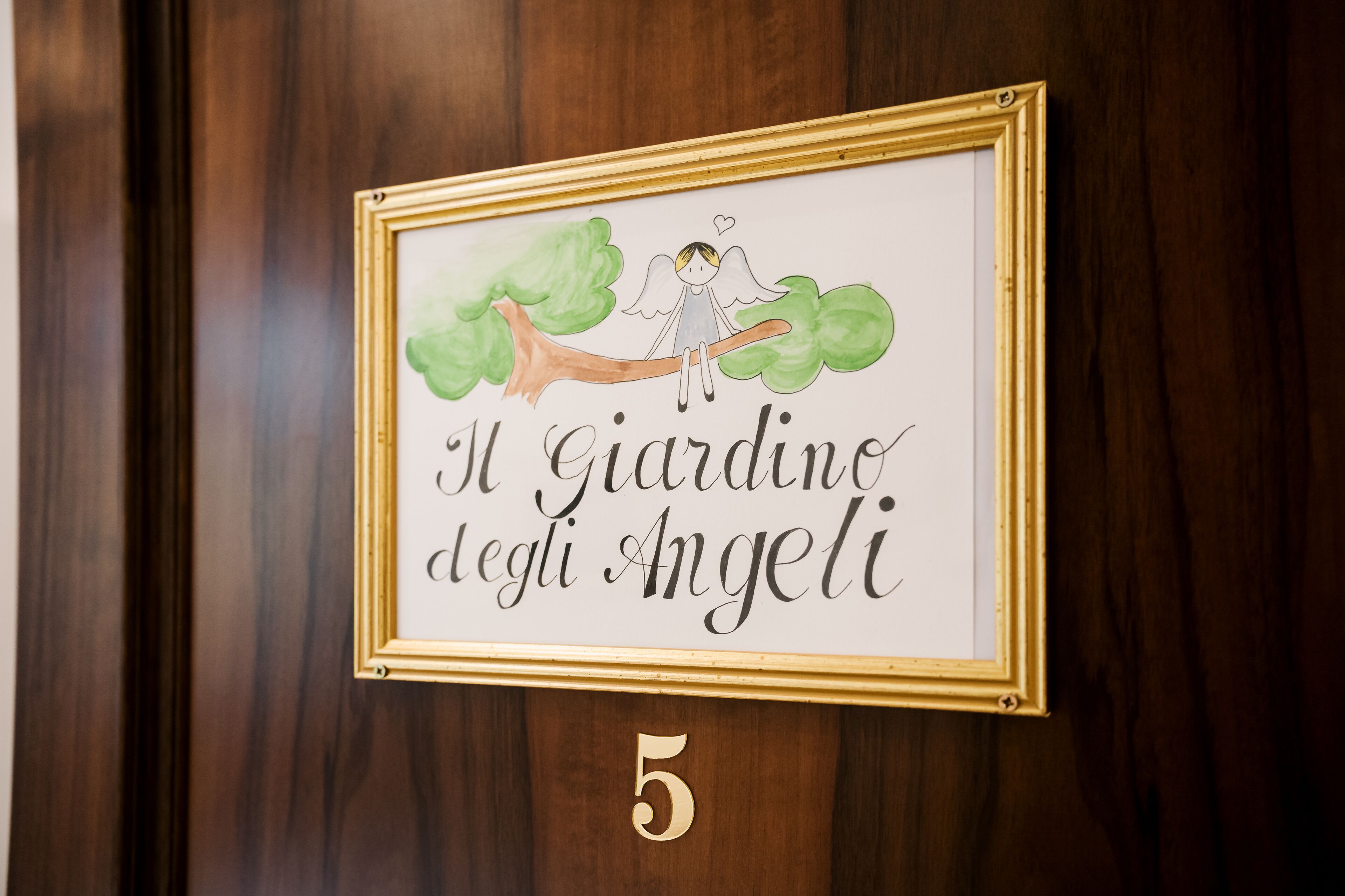 A framed sign with a drawing of an angel on a branch and the text "Il Giardino degli Angeli" on a wooden door.