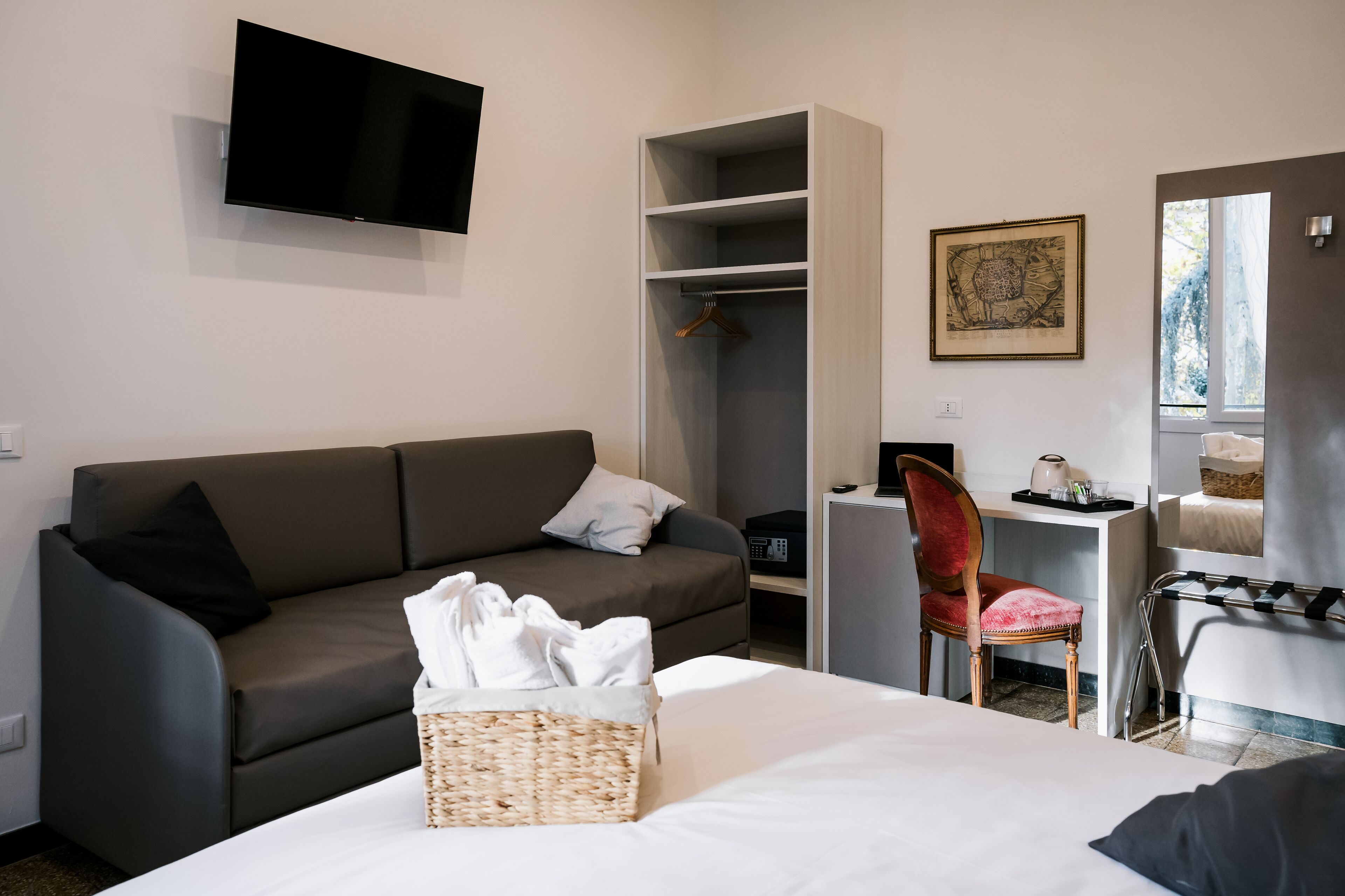 A modern hotel room with a grey sofa, a bed with a white comforter, a desk with a chair, a wardrobe, and a flat-screen TV mounted on the wall.