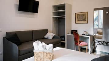 A modern hotel room with a grey sofa, a bed with a white comforter, a desk with a chair, a wardrobe, and a flat-screen TV mounted on the wall.