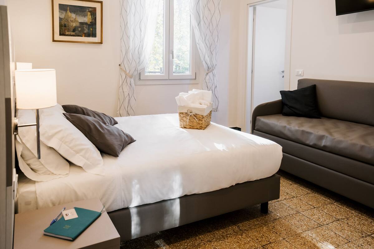 Un lit bien fait, avec une literie blanche et grise, une table de chevet avec une lampe et un carnet, un panier en osier rempli de serviettes et un canapé foncé.