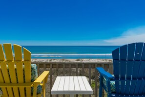 Une terrasse en bois avec deux fauteuils Adirondack et une petite table, donnant sur une plage sous un ciel bleu dégagé.