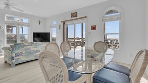 Amplia sala de estar con mesa de comedor de vidrio rodeada de sillas de mimbre, sofá con estampado colorido y televisor de pantalla plana.