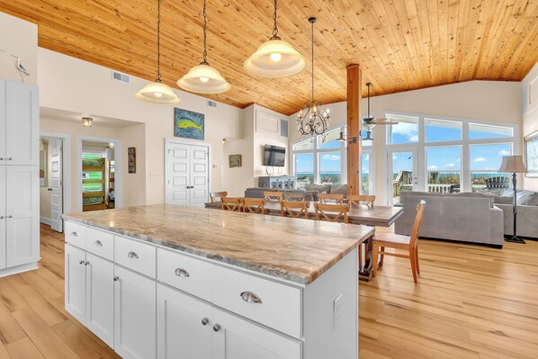 A spacious kitchen with a large island, wooden ceiling, and large windows offering a view.