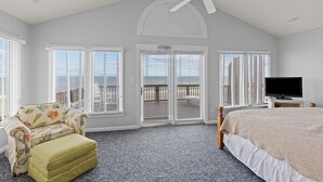 A bedroom with a bed, a chair, a television, and a balcony with white shutters. - 4BR Ocean Front - King MBR with Views! (Emerald Isle)