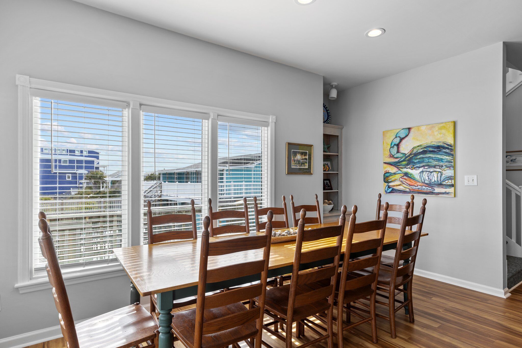 A dining area with a wooden table and chairs, large windows with blinds, a bookshelf, and a colorful painting on the wall.