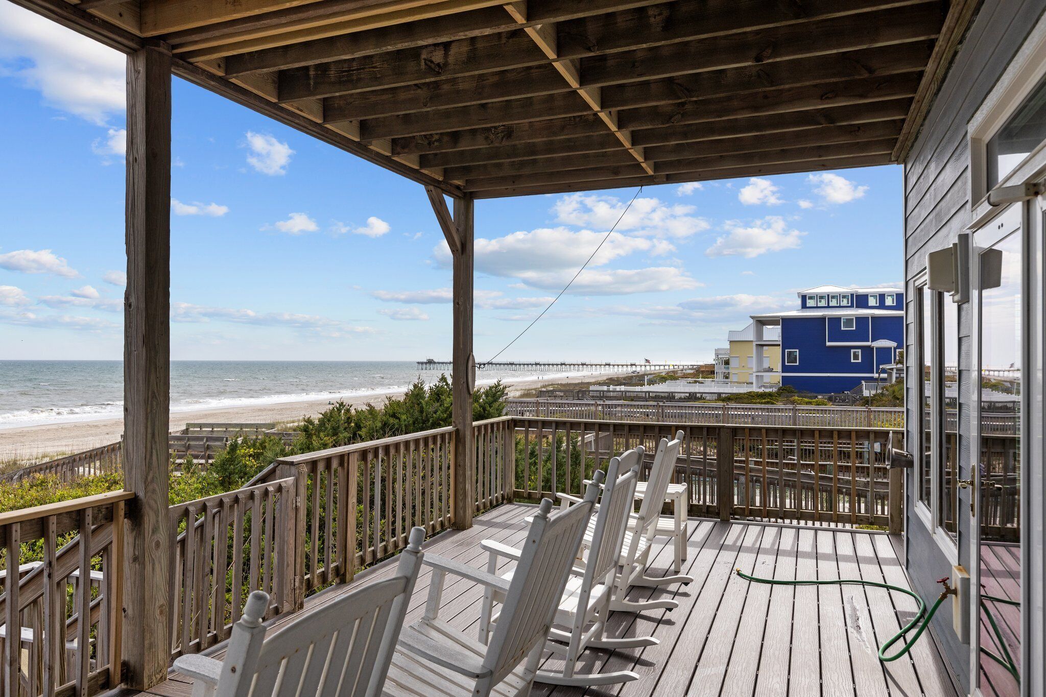A wooden deck with white rocking chairs overlooking a beach and a pier.