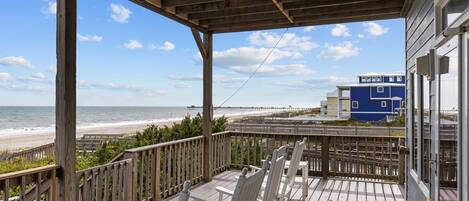 A wooden deck with white rocking chairs overlooking a beach and a pier.