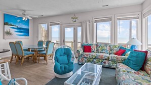 A spacious living room with a large window, a ceiling fan, a dining area with wicker chairs, and a colorful sofa. - 4BR Ocean Front Dog Friendly Duplex - Rent both sides! (Emerald Isle)