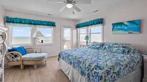 A bedroom with a large bed, a wicker chair, a nightstand, and a view of the ocean. - 4BR Ocean Front Dog Friendly Duplex - Rent both sides! (Emerald Isle)