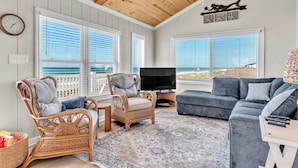 A room with a ceiling fan, a television, wicker furniture, a gray sofa, and large windows. - 4BR Ocean Front - Dogs OK! (Emerald Isle)