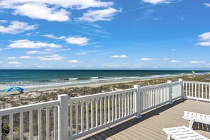 Eine Holzterrasse mit weißen Geländern, die auf einen Strand mit Wellen und blauem Himmel blickt.