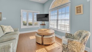 A living room with a sofa, a round table, a patterned armchair, and a TV mounted on the wall. - 4BR Ocean Front - DOG OK - Close to town! (Emerald Isle)
