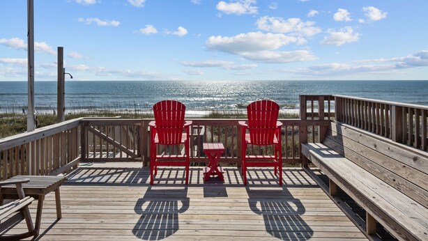 Une terrasse en bois avec deux chaises rouges et une petite table, donnant sur une plage, l’océan et un ciel dégagé.