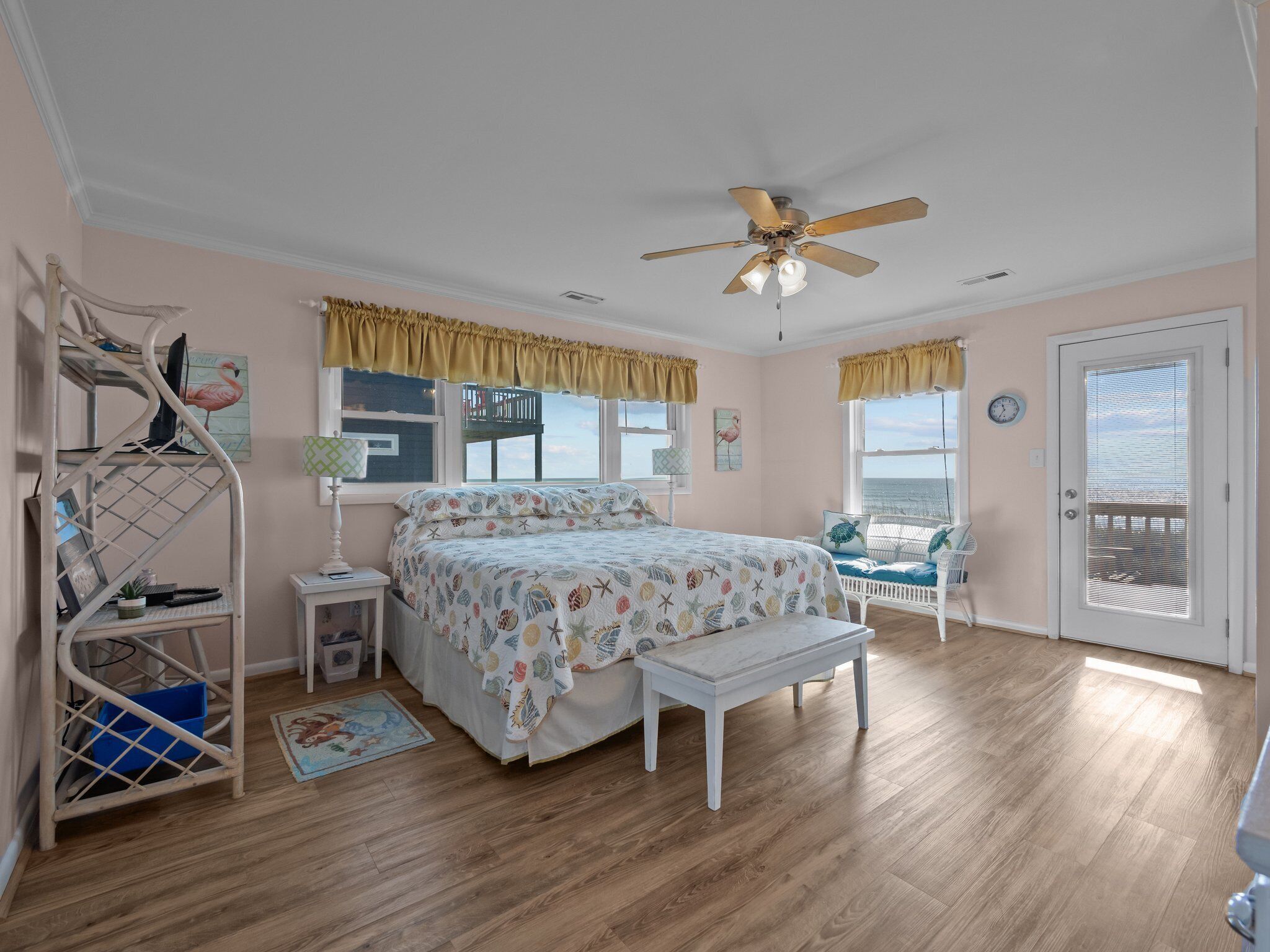 A bedroom with a bed, a ceiling fan, a desk, a chair, and a view of the ocean through a door.
