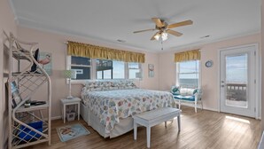 Une chambre à coucher comprenant un lit, un ventilateur de plafond, un bureau, une chaise et une vue sur l’océan, visible par la porte.
