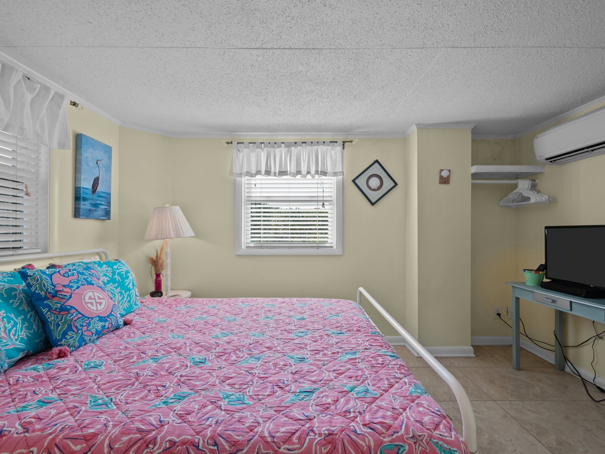 A bedroom with a bed, a window with blinds, a desk with a computer, and an air conditioning unit.