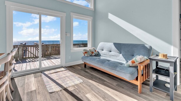 A spacious room with a large window overlooking a wooden deck and the ocean, a gray sofa with decorative pillows, and a small table with a book and a lamp. - 3BR - One Level - SUPER CUTE - Ocean front & DOGS OK!! (Emerald Isle)