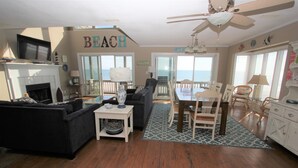 A living room with a fireplace, a television, a dining area with a table and chairs, and a view of the ocean through large windows. - 3BR- Dogs OK- Ocean Front - Master Suite (Emerald Isle)