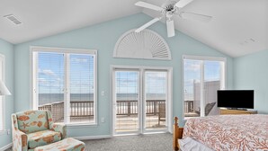 A bedroom with a bed, a chair, an ottoman, a TV, and a ceiling fan.