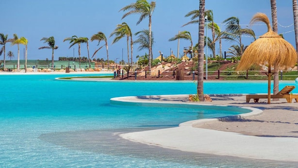 Ein Resort-Pool mit klarem türkisblauem Wasser, umgeben von Palmen und einem Reedschirm.