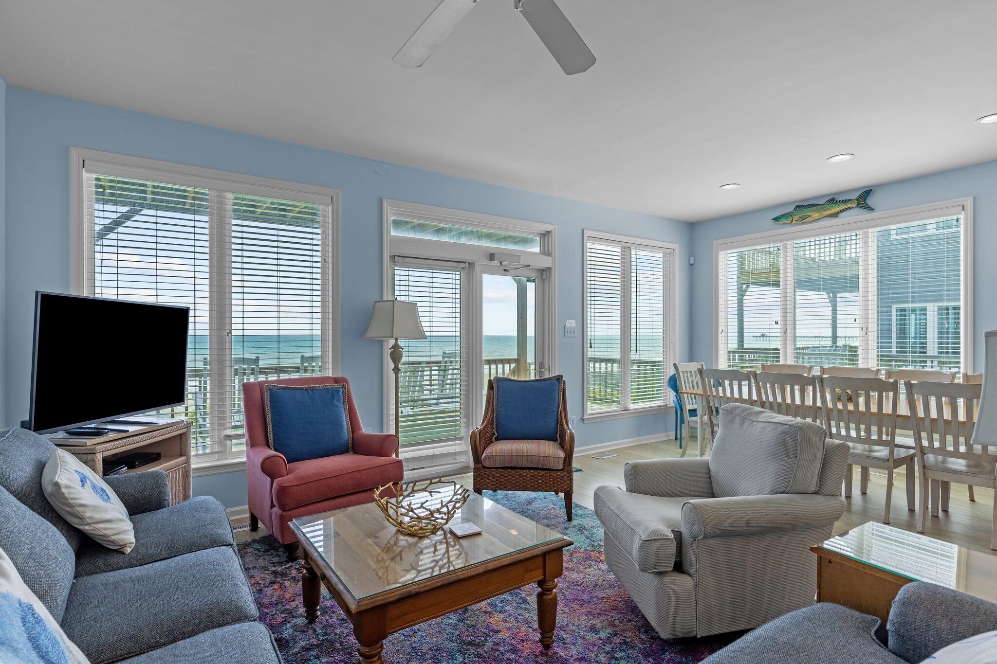 A spacious living room with a large TV, comfortable seating, and a view of the ocean through large windows.