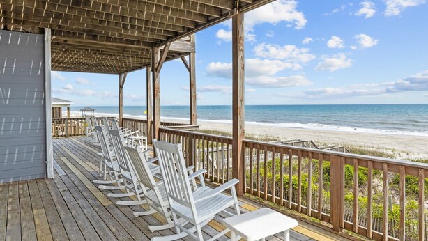 A wooden deck with white rocking chairs and a table, overlooking a beach with a wooden railing. - 4BR duplex close to town and ocean front! (Emerald Isle)