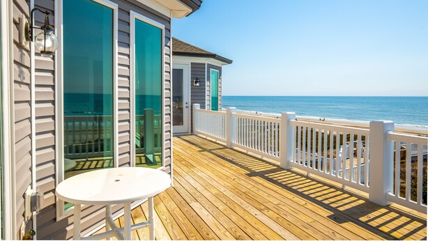 Une terrasse en bois avec une balustrade blanche, une porte vitrée et une vue sur l’océan.