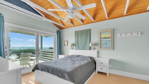 Une chambre à coucher avec un lit, un ventilateur de plafond, une table de chevet, une commode, une fenêtre donnant sur un beau paysage et un tableau accroché au mur.