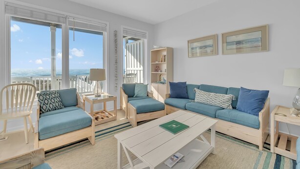 A modern living room with a view of the sea, featuring a blue sofa, a wooden coffee table, and a bookshelf. - 4BR Ocean Front Duplex - off Coast Guard Rd! (Emerald Isle)