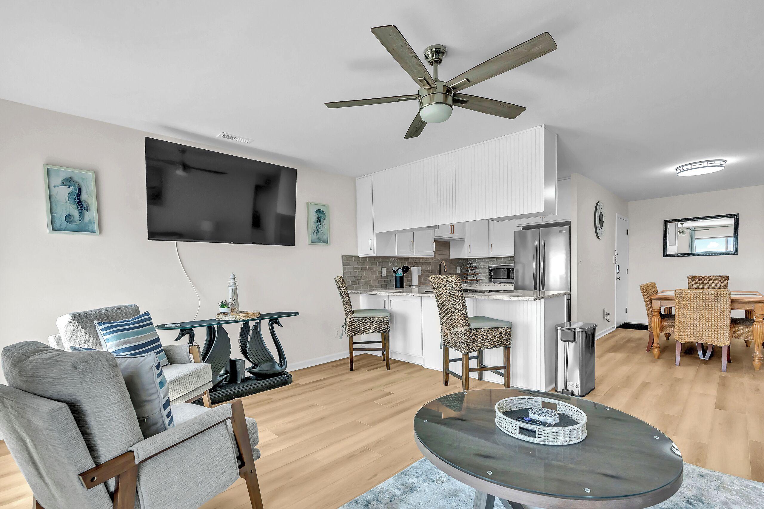 A modern living room with a glass coffee table, grey armchairs, a ceiling fan, a kitchen area with white cabinets, and a dining area with wicker chairs.
