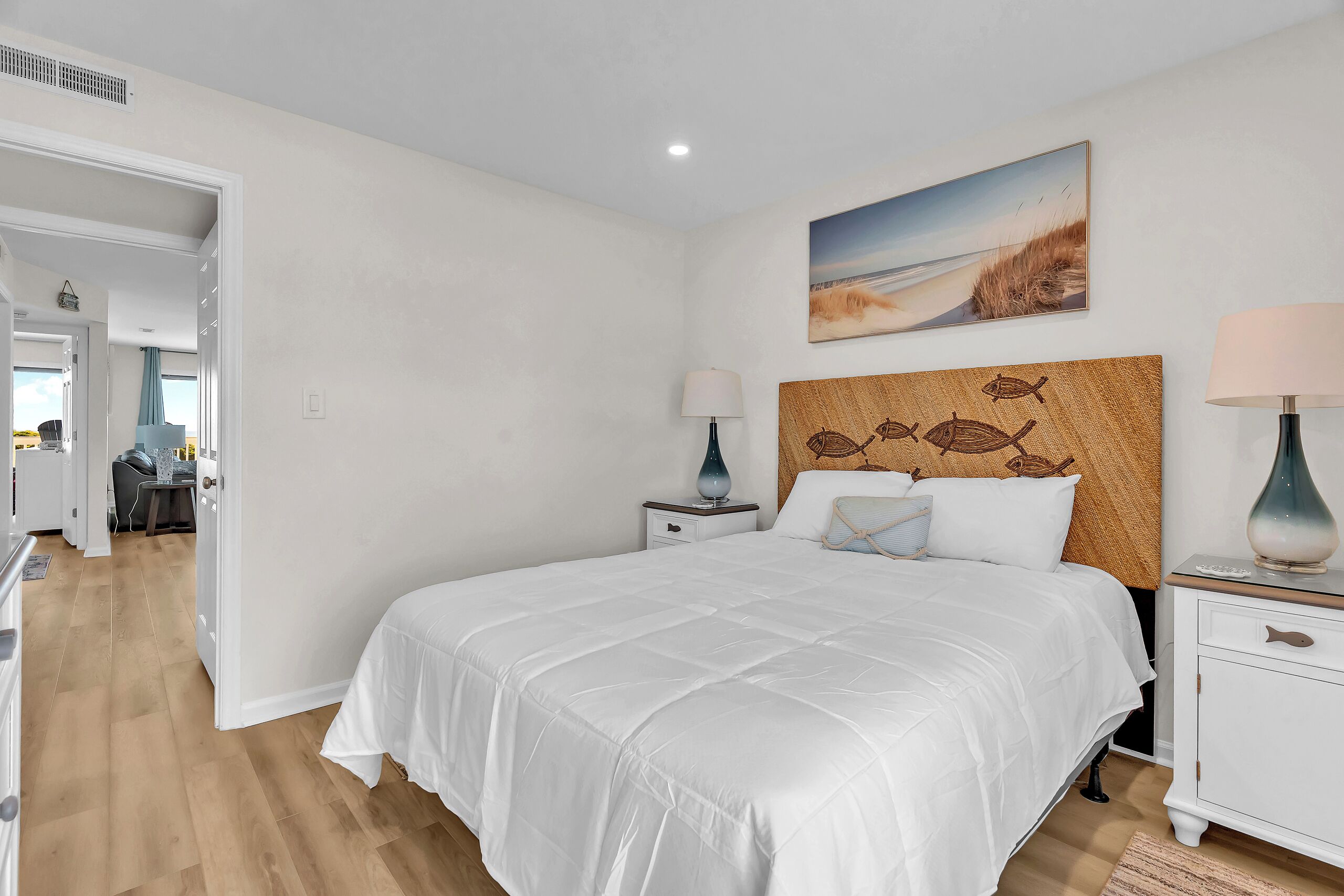 A bedroom with a large bed, a headboard with fish designs, a nightstand with lamps, and a painting of a beach scene.