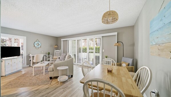 A spacious living area with a sofa, dining table, and chairs, a television, and a large window with blinds.