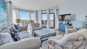 A modern living room with a sofa, ottoman, and armchairs, a flat-screen TV, and a balcony with a view. - Point Emerald E305 - 3BR - Near "The Point" and views of Bogue Inlet (Emerald Isle)