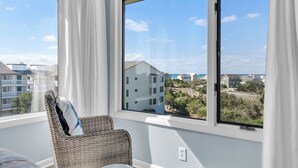 A room with a large window, wicker chair, and a view of buildings and trees. - Point Emerald E305 - 3BR - Near "The Point" and views of Bogue Inlet (Emerald Isle)