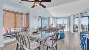 A spacious living area with a glass dining table, comfortable seating, a ceiling fan, and large windows offering a view. - Point Emerald E305 - 3BR - Near "The Point" and views of Bogue Inlet (Emerald Isle)