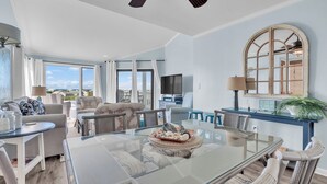 A spacious living area with a glass dining table, comfortable seating, a large mirror, and a view of the outdoors. - Point Emerald E305 - 3BR - Near "The Point" and views of Bogue Inlet (Emerald Isle)