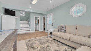 A modern living room with a beige sofa, a patterned rug, a flat-screen TV, and a staircase leading to a second floor. - Pool & Hot Tub- DOGS OK! 5BR w/ Ocean Views! (Emerald Isle)