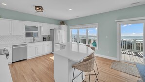 A modern kitchen with white cabinets, a central island, and a view of the ocean through large windows. - Pool & Hot Tub- DOGS OK! 5BR w/ Ocean Views! (Emerald Isle)