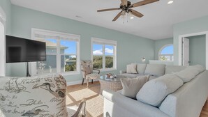 A spacious living room with a ceiling fan, a flat-screen TV, and a large window offering a view of the outdoors. - Pool & Hot Tub- DOGS OK! 5BR w/ Ocean Views! (Emerald Isle)